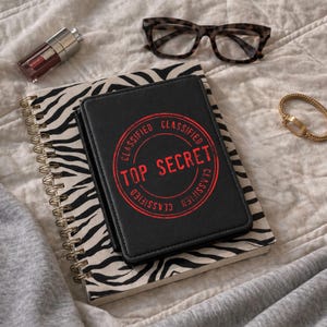 May include: A black notebook with the words "TOP SECRET" in red, stamped on the cover, rests on a zebra-print spiral notebook. A pair of eyeglasses, a gold bracelet, and a tube of lipstick are also visible.
