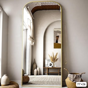 May include: A tall, arched-top mirror with a gold-colored frame. The mirror is 71 inches tall and 28 inches wide. The reflection shows a room with neutral tones, a window, and decorative items. The mirror is a decorative home decor item.