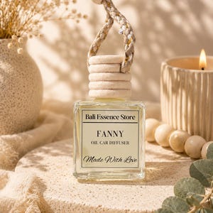 May include: A square glass car oil diffuser bottle with a light-colored liquid inside. The label reads Bali Essence Store, FANNY, OIL CAR DIFFUSER, and Made With Love. It features a light wooden cap and a braided cord with a wooden bead for hanging.