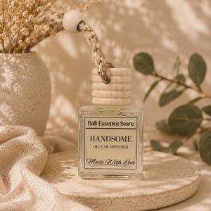 Puede incluir: Un frasco difusor de aceite para coche de vidrio cuadrado con tapa de madera clara y cordón trenzado para colgar. La etiqueta muestra el texto Bali Essence Store, HANDSOME, OIL CAR DIFFUSER y Made With Love en negro sobre fondo crema.