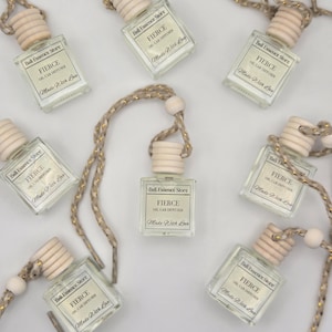 Puede incluir: Vista cenital de varios ambientadores para coche de cristal transparente. Cada botella tiene una tapa de madera y está suspendida por una cuerda beige y dorada. La etiqueta dice "FIERCE OIL CAR DIFFUSER Made With Love" de Bali Essence Store.