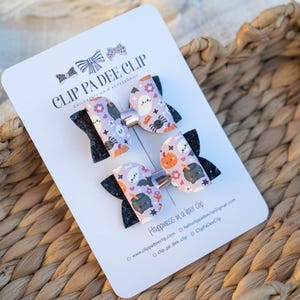 May include: Two Halloween hair bows on a white card with the text "CLIP PA DEE CLIP". The bows have a black glitter base and a patterned top with ghosts, pumpkins, and spiders. The card rests on a woven basket.