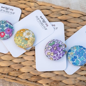 May include: Four floral fabric hair clips on white cards, arranged on a woven surface. Each clip features a different colourful floral pattern, including red, purple, yellow, and blue. The cards have the text "CLIP PA DEE CLIP".