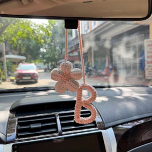 Op de afbeelding: Een handgemaakte, lichtroze gehaakte auto-hanger. De hanger heeft een bloem en de letter "B". De hanger hangt aan de achteruitkijkspiegel van een auto. Het dashboard van de auto is zichtbaar op de voorgrond.