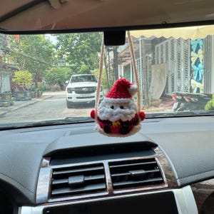 May include: A crocheted Santa Claus ornament with a red hat, white beard, and black and red suit hangs from a car's rearview mirror. The ornament is suspended by a cream-colored string, adding a festive touch to the vehicle's interior.