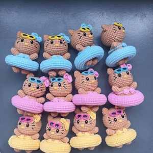 May include: A collection of crocheted cat figurines. Each cat is wearing sunglasses and a crocheted floatie. The floaties are blue, pink, and yellow. The cats have brown bodies, black whiskers, and are wearing sunglasses.