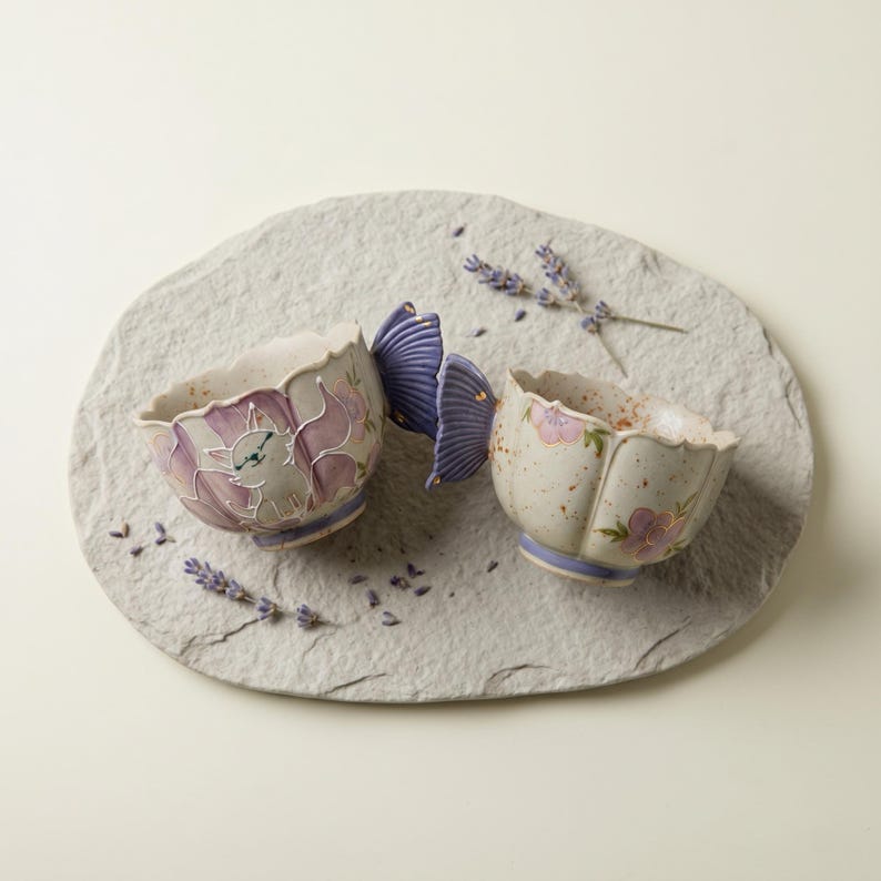 May include: Two ceramic bowls with butterfly wing handles, resting on a gray stone. The bowls are beige with purple floral and cat designs. Lavender sprigs are scattered around the bowls, creating a serene, nature-inspired aesthetic.