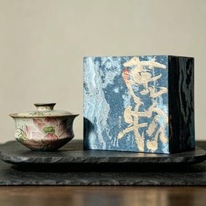 May include: A ceramic tea bowl with a lid featuring a hand-painted pink lotus flower design, positioned next to a rectangular blue box with a marbled texture and gold-colored calligraphic text printed on the front surface.