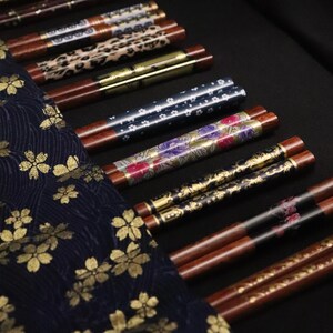 Könnte beinhalten: Eine Auswahl an dekorativen Essstäbchen mit verschiedenen Designs, darunter Blumenmuster, Leopardenmuster und metallische Akzente. Die Essstäbchen werden auf einer schwarzen Oberfläche präsentiert, mit einem marineblauen Stoff mit goldenen Blumenmustern im Vordergrund.