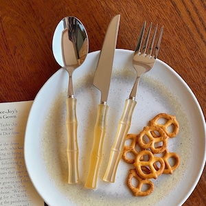 May include: A stainless steel spoon, knife, and fork set with bamboo-style handles, arranged on a speckled white plate with pretzels. The handles are a light amber color. The spoon reflects the light.