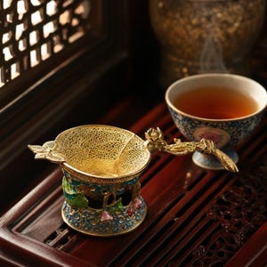 Puede incluir: Un colador de té dorado con un diseño floral ornamentado, colocado sobre una base decorativa con detalles azules, verdes y rosas. El colador está junto a una taza de té llena de té.