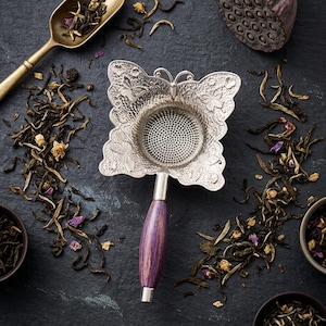 Puede incluir: Un infusor de té plateado con forma de mariposa y mango de madera púrpura, rodeado de hojas de té sueltas y una cuchara dorada. El infusor tiene detalles florales intrincados y un filtro de malla fina. Las hojas de té son verdes y marrones, con flores secas.