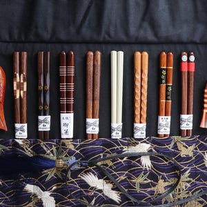 May include: A collection of wooden chopsticks in various designs, including carved and patterned styles, displayed with matching fish-shaped chopstick rests. The chopsticks are presented on a black surface with a patterned fabric.