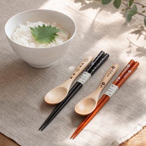 Könnte beinhalten: Zwei Sets aus Holzstäbchen gepaart mit passenden Holzlöffeln. Ein Set ist schwarz mit Vogelmotiven, das andere rotbraun mit Blumenmustern. Jeder Löffelstiel zeigt eine kleine Grafik, und ein Papierband mit der Aufschrift Natural wood ist um jedes Paar gewickelt.