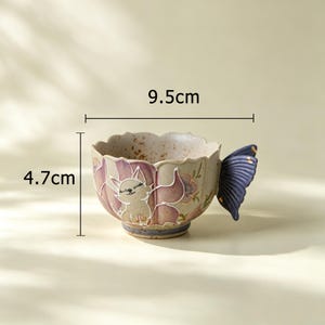 May include: A ceramic teacup with a fox design and a butterfly-shaped handle. The cup is 3.7 inches wide and 1.9 inches tall. The cup has a beige base with a purple and white fox illustration and purple floral accents.