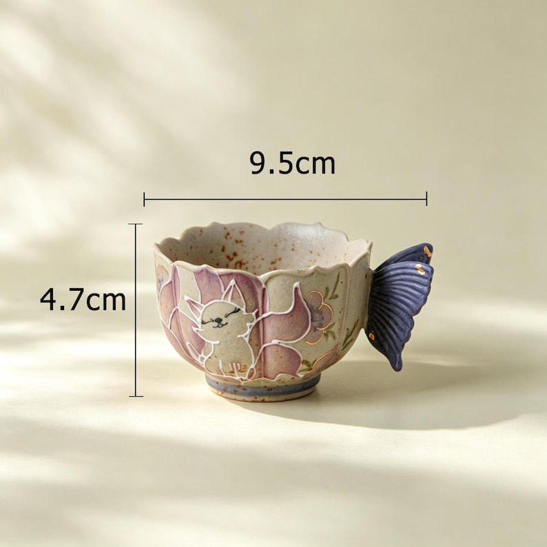 May include: A ceramic teacup with a fox design and a butterfly-shaped handle. The cup is 3.7 inches wide and 1.9 inches tall. The cup has a beige base with a purple and white fox illustration and purple floral accents.