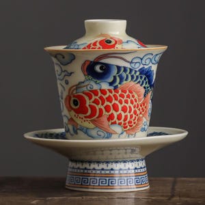 May include: A ceramic tea set featuring a cup, lid, and saucer, all resting on a decorative stand. The cup and lid are adorned with vibrant illustrations of red and blue koi fish swimming amidst clouds. The stand has a patterned design.