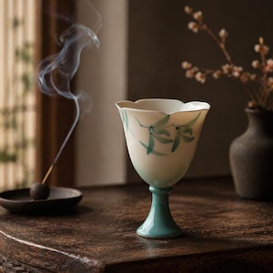 May include: A ceramic goblet with a scalloped rim and a flared base. The cup features a white body with a soft green gradient near the stem and hand-painted green leaf motifs. The stem is a solid, glossy teal color, contrasting with the cup.