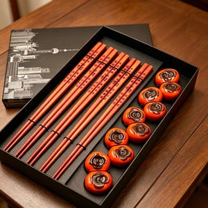May include: A set of five pairs of reddish-brown wooden chopsticks with black Chinese characters, accompanied by ten small, round, orange-colored ceramic chopstick rests shaped like persimmons, all presented in a black rectangular gift box.