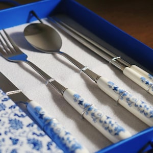 Puede incluir: Un juego de cubiertos y palillos chinos de acero inoxidable con mangos de porcelana blanca con diseños florales azules. El juego incluye un tenedor, cuchillo, cuchara y palillos, presentado en una caja azul.