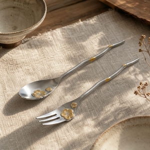 May include: A silver-toned metal spoon and fork set featuring a textured, branch-like handle design. Each piece is adorned with small, raised gold-colored flower accents on the handle and the bowl of the spoon and the base of the fork tines.
