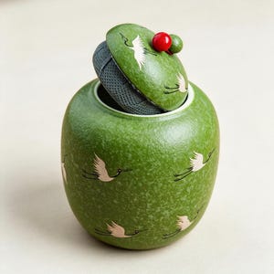 May include: A round green ceramic tea canister with a textured finish, decorated with white crane illustrations. The lid features a red and green spherical knob and is slightly ajar, revealing a dark grey mesh tea strainer inside the container.