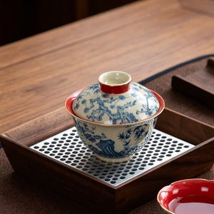 Op de afbeelding: Een keramische gaiwan theekom met deksel, voorzien van een handgeschilderd blauw-wit landschapspatroon van bomen en rotsen. De randen van de kom en het deksel zijn geaccentueerd met een dunne gouden rand en het deksel heeft een rode knop.