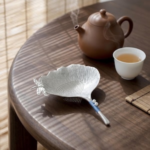 May include: A silver tea strainer with a dragonfly accent, a brown teapot, and a white teacup filled with tea on a wooden table. Steam rises from the teapot, suggesting freshly brewed tea. A bamboo mat is also present.