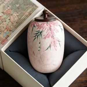 May include: A cylindrical ceramic tea canister with a soft pink mottled finish, featuring hand-painted pink floral clusters and green foliage. The container is topped with a dark brown wooden lid with a small knob handle, presented in a box.