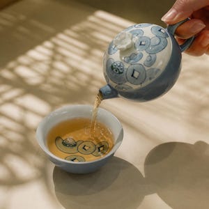 Peut inclure: Une théière en céramique bleu clair avec un couvercle blanc orné d'un bouton floral et de motifs de pièces bleues, versant du thé ambré dans une tasse assortie présentant un motif de pièce peint au fond de son bassin intérieur.