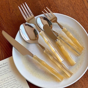 Puede incluir: Un juego de cubiertos de acero inoxidable con mangos amarillos estilo bambú, que incluye tenedores, cucharas y cuchillos, dispuestos sobre un plato blanco. Los cubiertos son reflectantes y los mangos tienen un aspecto natural y texturizado. Un recorte de revista es visible.