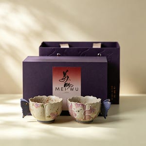 May include: Two ceramic teacups with butterfly wing handles and floral and cat designs. The cups are cream-colored with pink and purple accents. They are presented in front of a purple gift box with the text "MEI WU" on the front.
