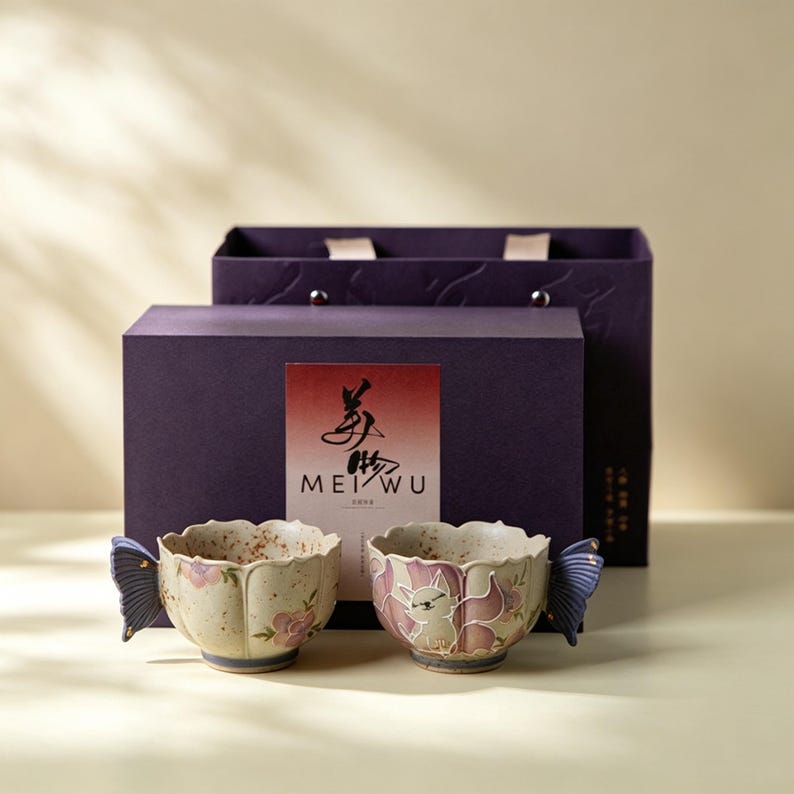 May include: Two ceramic teacups with butterfly wing handles and floral and cat designs. The cups are cream-colored with pink and purple accents. They are presented in front of a purple gift box with the text "MEI WU" on the front.