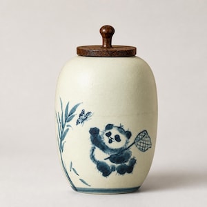 May include: A cream-colored ceramic jar with a dark brown wooden lid featuring a knob handle. The jar is decorated with a blue painted design of a panda holding a butterfly net, a small butterfly, and bamboo stalks on the side of the vessel.