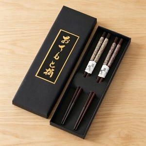 May include: A set of two pairs of dark brown chopsticks presented in a black rectangular gift box. The upper sections of the chopsticks feature textured, speckled inlays in shades of gray and brown. The box lid displays gold Japanese text.