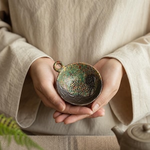 May include: A round ceramic tea strainer with a small handle, featuring a speckled green and brown glaze. The interior base has a perforated mesh area for straining and a small embossed lotus pod design on the side with a dark brown rim.