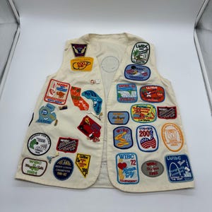 May include: Cream-colored vest covered in colorful patches, many with the letters "WIBC" and various city names. Patches feature images of states, landmarks, and text. The vest has a simple, sleeveless design.
