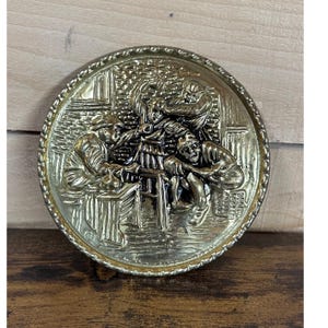 May include: A circular brass wall plaque featuring a high-relief embossed scene of three figures gathered around a table. The metallic surface has a polished gold-tone finish with darkened recessed areas to highlight the intricate details of the figures.