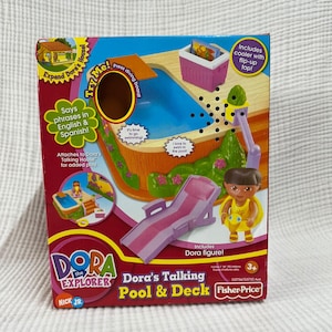 May include: Dora the Explorer Talking Pool & Deck playset in a box. The box is red and features a pool, deck, lounge chair, Dora figure, and a cooler. The playset says phrases in English and Spanish. Includes a diving board and a cooler with a flip-up top.