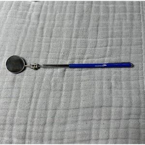 May include: A round, chrome inspection mirror with a blue handle. The handle has the word "MUSTANG" printed on it. The mirror is extended and lies on a white, textured surface.