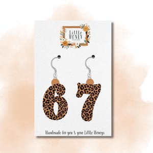 May include: A pair of earrings shaped like the numbers 6 and 7, featuring a brown and black leopard print pattern. The earrings hang from silver hooks with a small wooden bead. The card reads "Handmade for you & your Little Honeys."