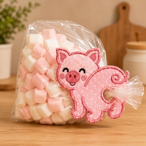 May include: A clear plastic bag filled with small, square, white and light pink marshmallows. Attached to the front of the bag is a pink, polka-dotted fabric pig cutout with embroidered facial features and a curled tail, featuring a circular cutout.