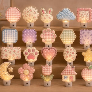 Puede incluir: Una colección de veinte luces nocturnas enchufables iluminadas con diversas formas de tela, incluyendo círculos, cuadrados, corazones, estrellas, flores, una luna, una casa, una manzana, una huella y un conejo, con suaves patrones pastel.