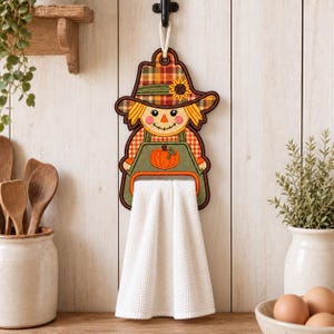 Puede incluir: Un soporte para toallas de cocina con forma de espantapájaros que presenta un sombrero de cuadros con un girasol, un delantal verde con un gráfico de calabaza naranja y una toalla blanca de textura tipo gofre colgada de un cordón.