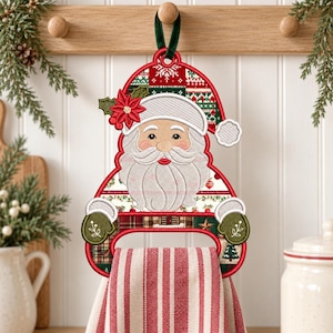 May include: A hanging kitchen towel holder featuring an embroidered Santa Claus design with a red hat, white beard, and green mittens. The holder includes a red and white striped fabric towel hanging from the centre, accented with festive patterns.