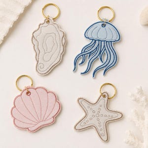 May include: Four embroidered keychains featuring marine life designs: a beige oyster shell, a light blue jellyfish with long tentacles, a pale pink scallop shell, and a beige starfish with dotted texture, each attached to a gold-toned metal ring.