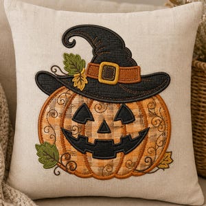 May include: A square beige throw pillow featuring an embroidered jack-o'-lantern wearing a black witch hat. The pumpkin has a plaid orange and tan pattern with black stitched eyes and a wide grin. Green leaves accent the base and hat brim.