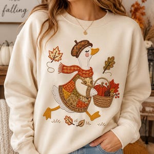 May include: A cream-colored crewneck sweatshirt featuring an embroidered graphic of a white duck wearing a brown beret and a plaid scarf. The duck is carrying a woven basket filled with a pumpkin, pinecone, and autumn leaves, surrounded by falling leaves.