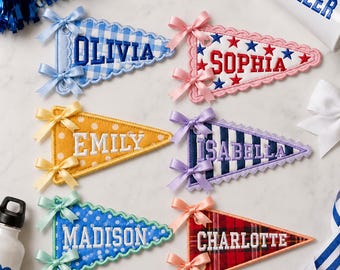 May include: Six triangular fabric pennants featuring embroidered names: Olivia, Sophia, Emily, Isabella, Madison, and Charlotte. Each pennant has scalloped edges, two satin ribbon bows, and distinct patterns including gingham, stars, polka dots, stripes, and plaid.