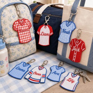 May include: A collection of personalized jersey-shaped keychains featuring various colors and patterns, including red gingham, pinstripes, and stars. Each keychain displays a name such as Mason, Liam, Noah, Jack, Emma, Owen, Ava, or Lily.